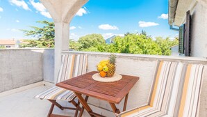 Ein Balkon mit einem Holztisch und Stühlen, einer gewebten Tischdecke mit Obst und Blick auf Grünflächen und Gebäude.