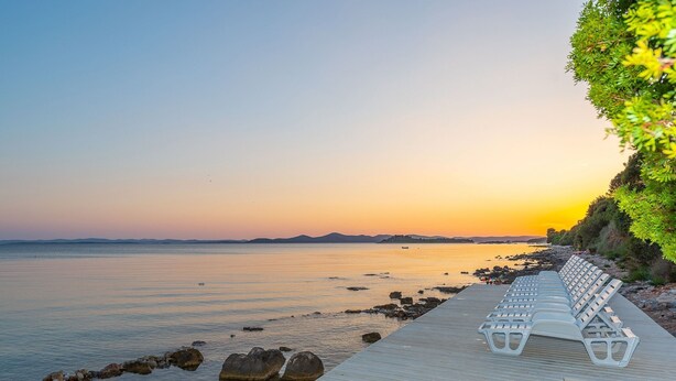 A wooden pier with white lounge chairs extending over calm water, surrounded by rocks and greenery. - Mobile Home Pecten 01 (Pakostane)