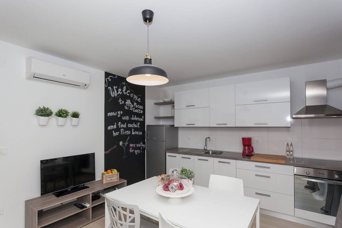 A modern kitchen with white cabinets, a black pendant light, a flat-screen TV, and a chalkboard with writing.