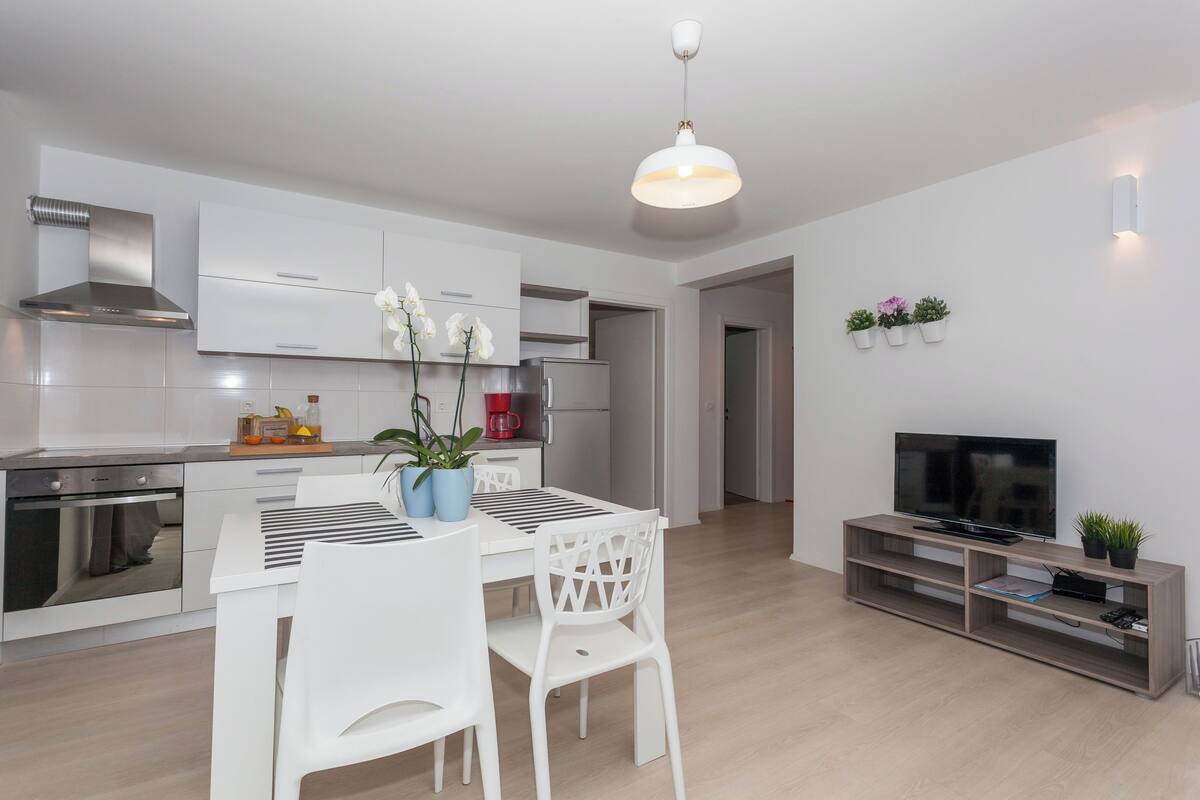 A modern kitchen with white cabinetry, a built-in oven, and a stainless steel range hood. There is a dining area with a white table and chairs, and a TV on a wooden stand.
