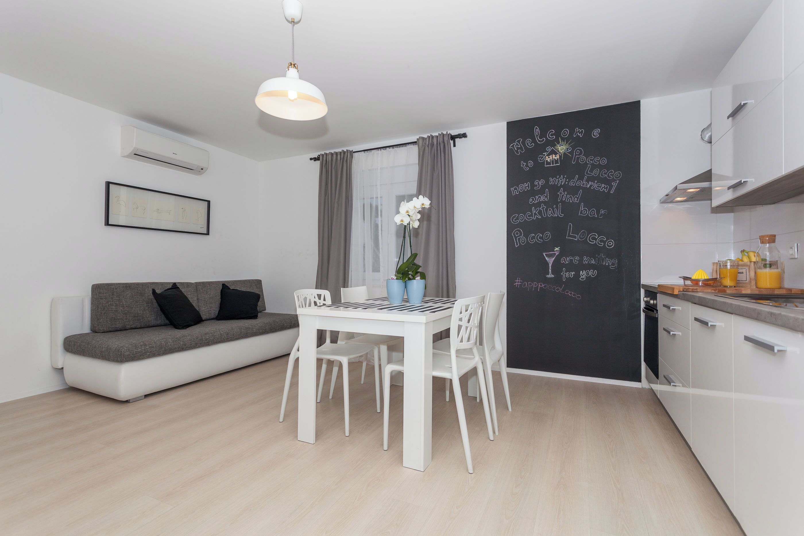 Eine moderne Küche mit weißen Schränken, einem Esstisch mit Stühlen, einem grauen Sofa und einer Tafel mit Schrift.
