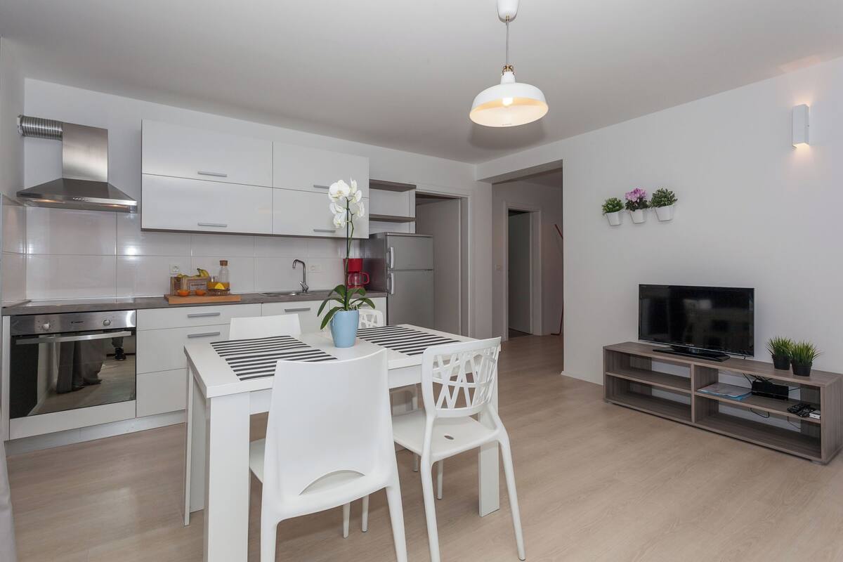 A modern kitchen with white cabinetry, a built-in oven, and a dining area with a white table and chairs.