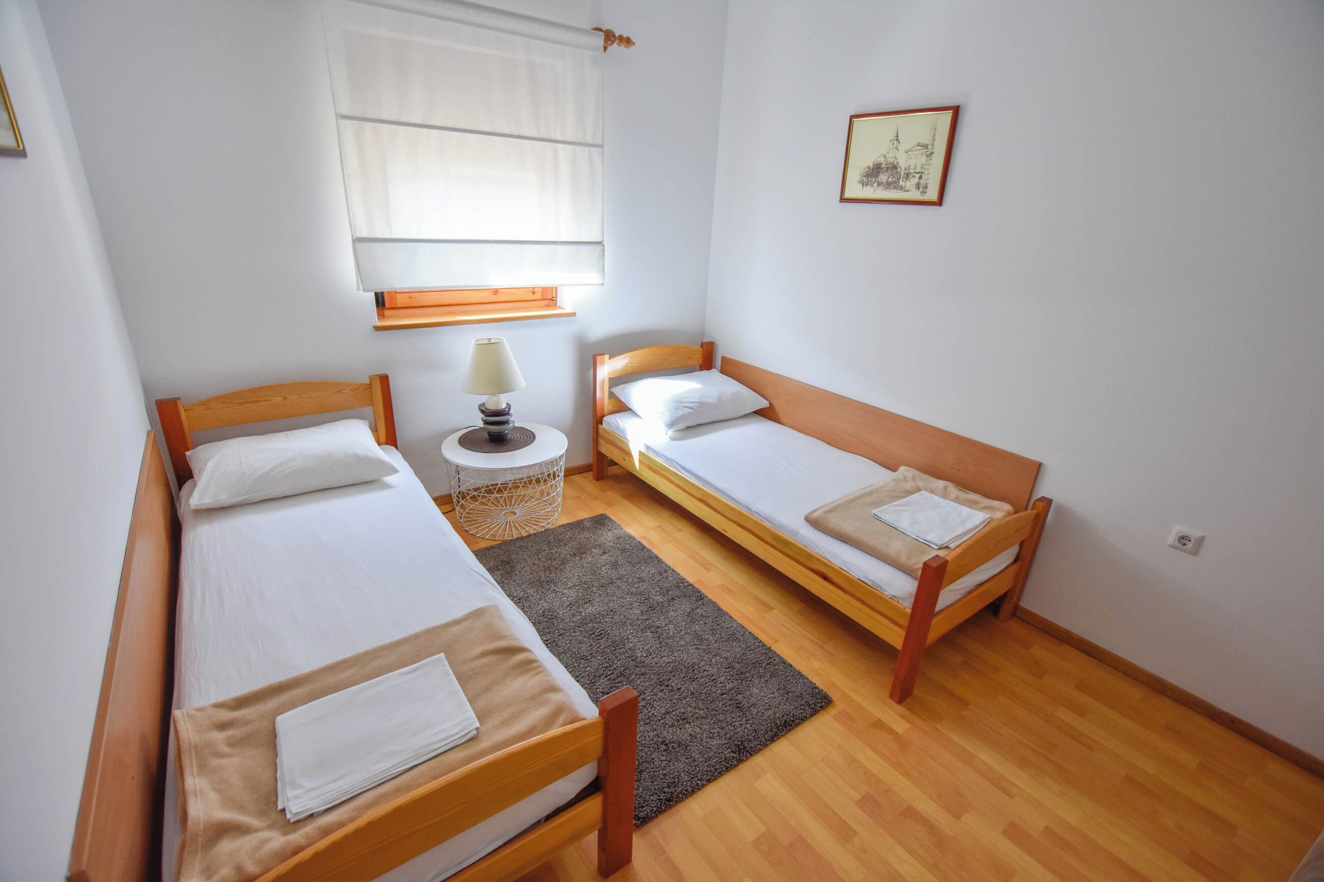Una habitación pequeña y limpia con dos camas de madera, una mesita redonda con una lámpara, una ventana con persianas y un cuadro en la pared.