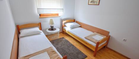 Ein kleiner, sauberer Raum mit zwei hölzernen Betten, einem runden Tisch mit Lampe, einem Fenster mit Jalousien und einem gerahmten Bild an der Wand.