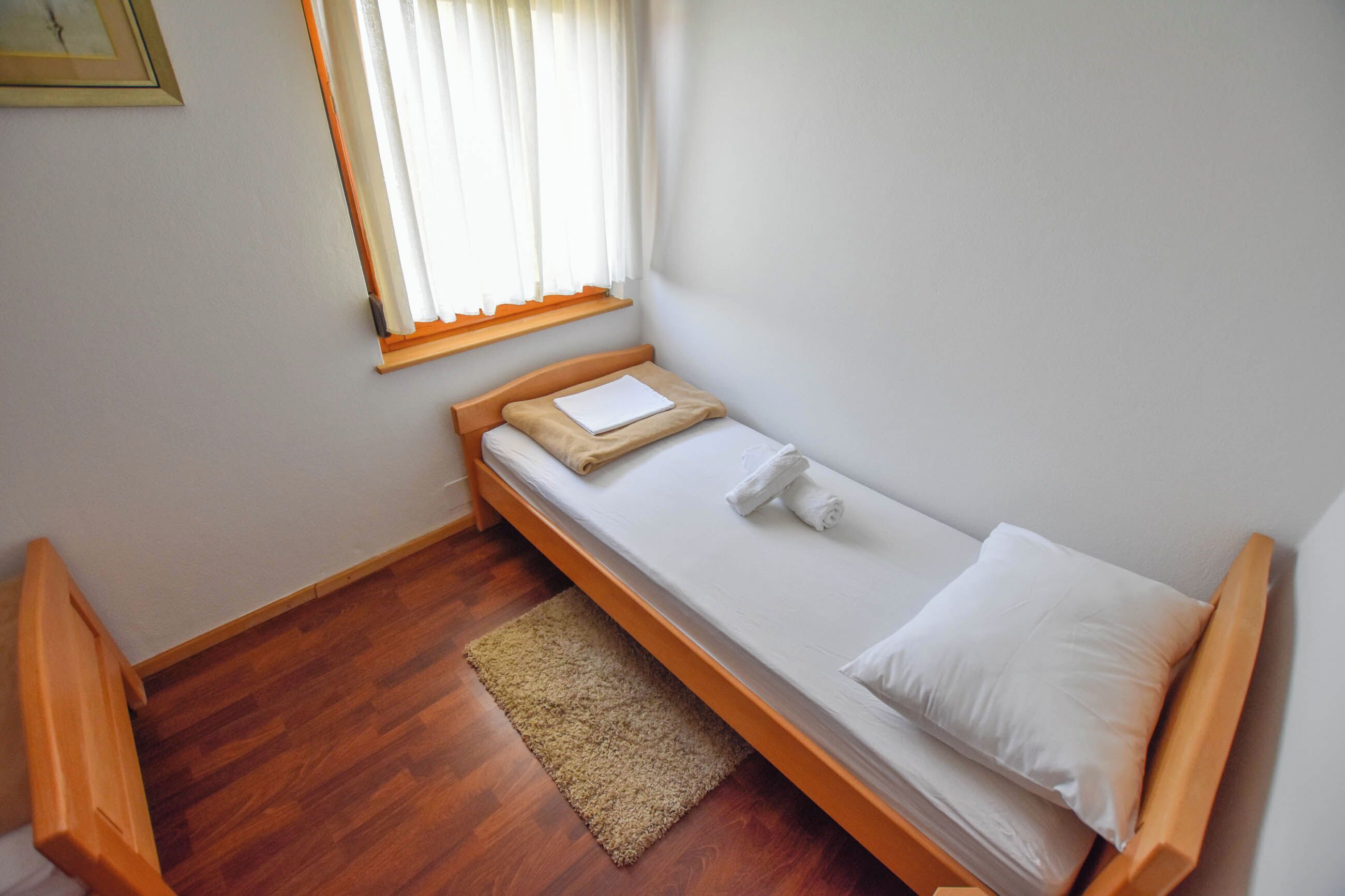 A small, single-bed room with a wooden frame, a window with blinds, a wooden floor, and a small rug.