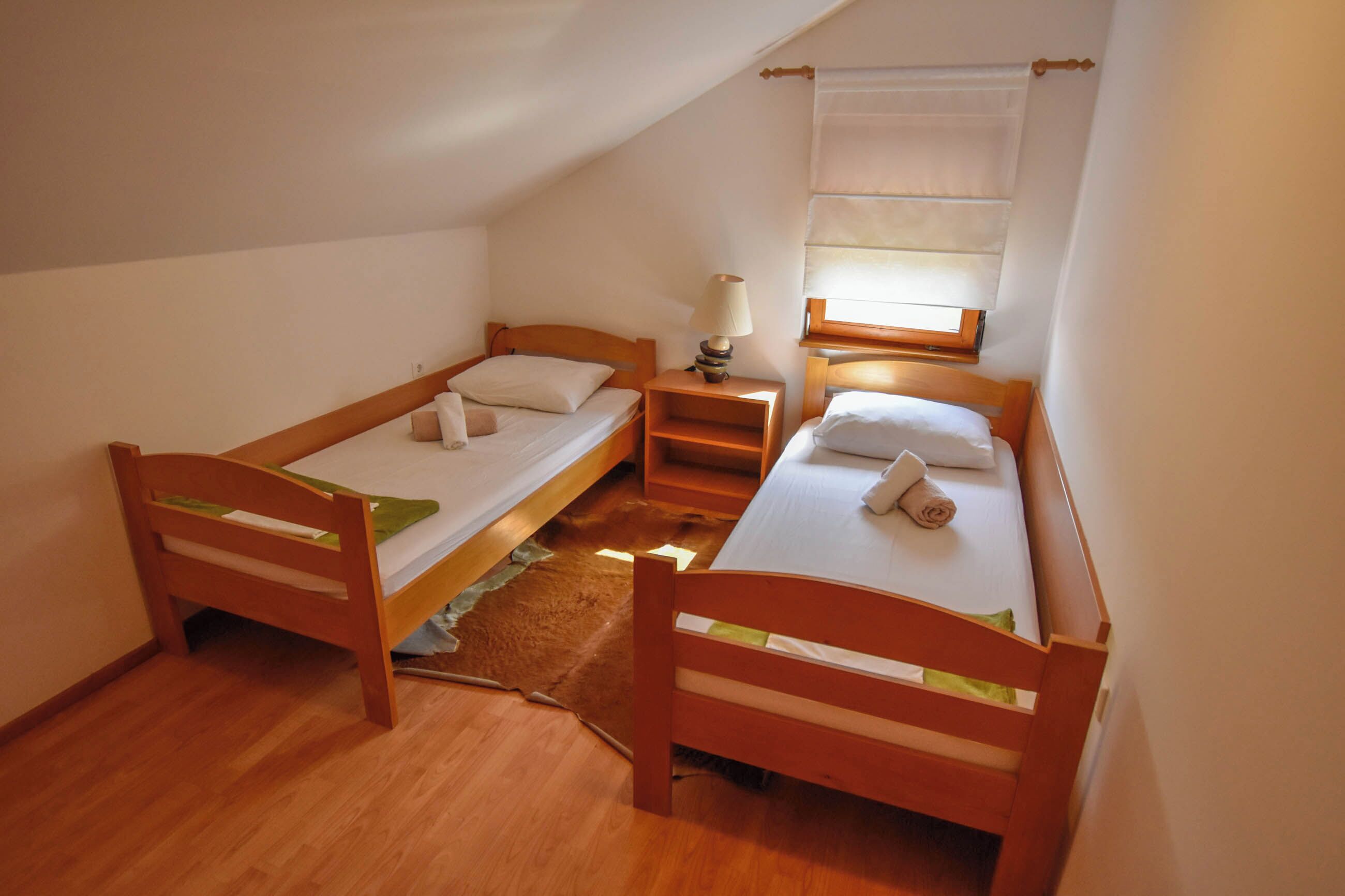 A small, cozy room with two wooden beds, a nightstand, a lamp, and a window with a curtain.