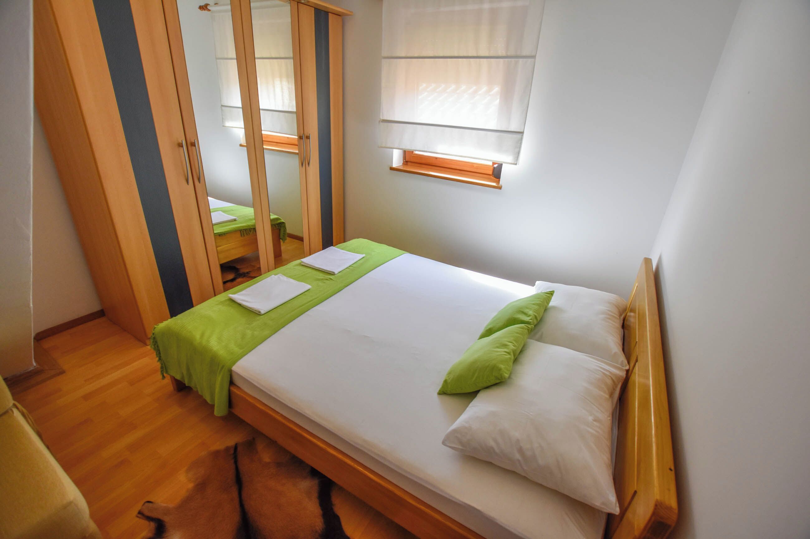 A small, neatly arranged bedroom with a single bed, a wooden wardrobe, a mirror, and a window with blinds.