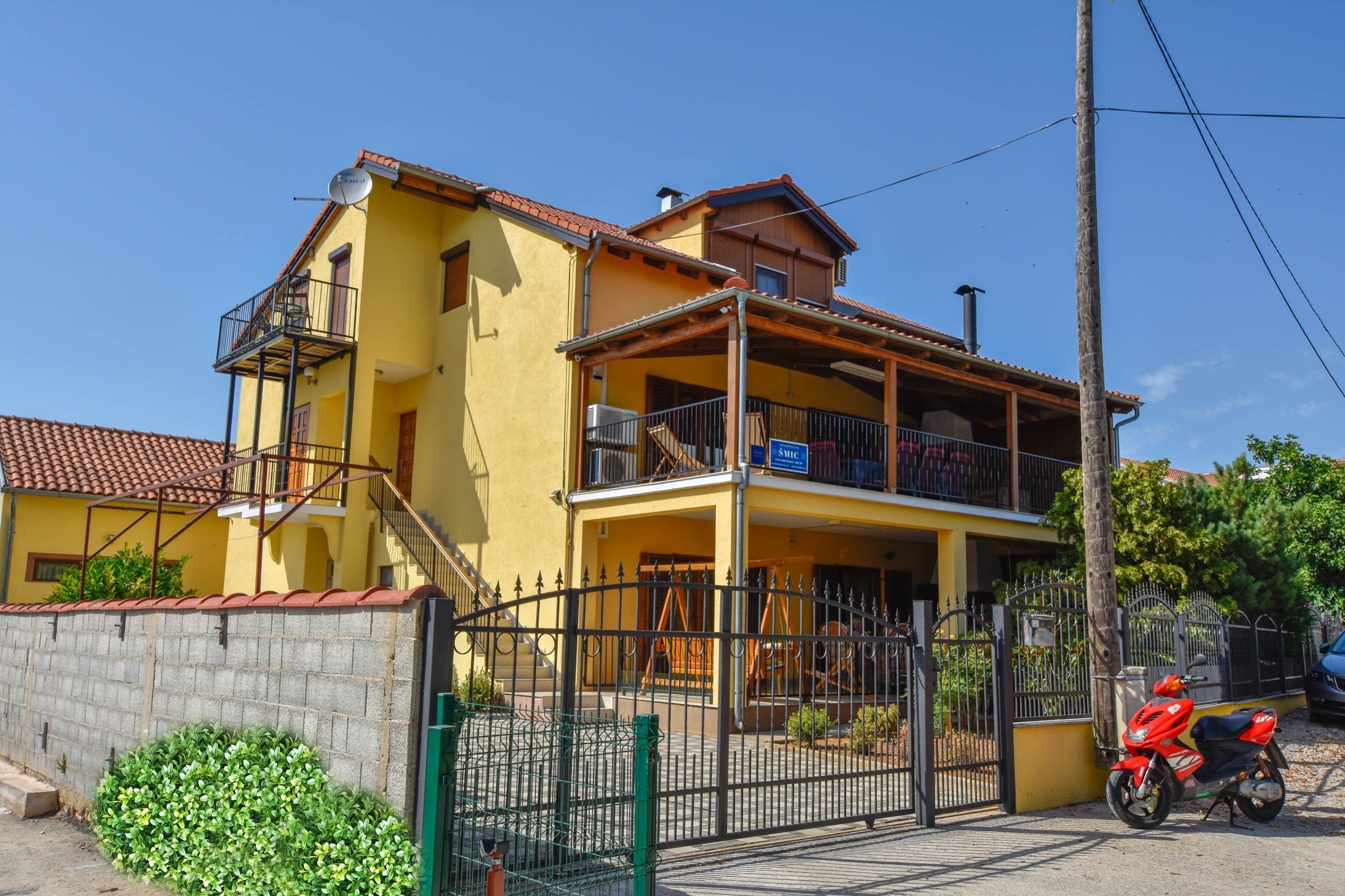 A two-story yellow building with a balcony, a gated entrance, and a red scooter parked outside.