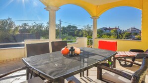 A covered outdoor patio with a table and chairs, a red cushioned bench, and a view of greenery and houses in the background. - Apartments Svarog (Biograd)