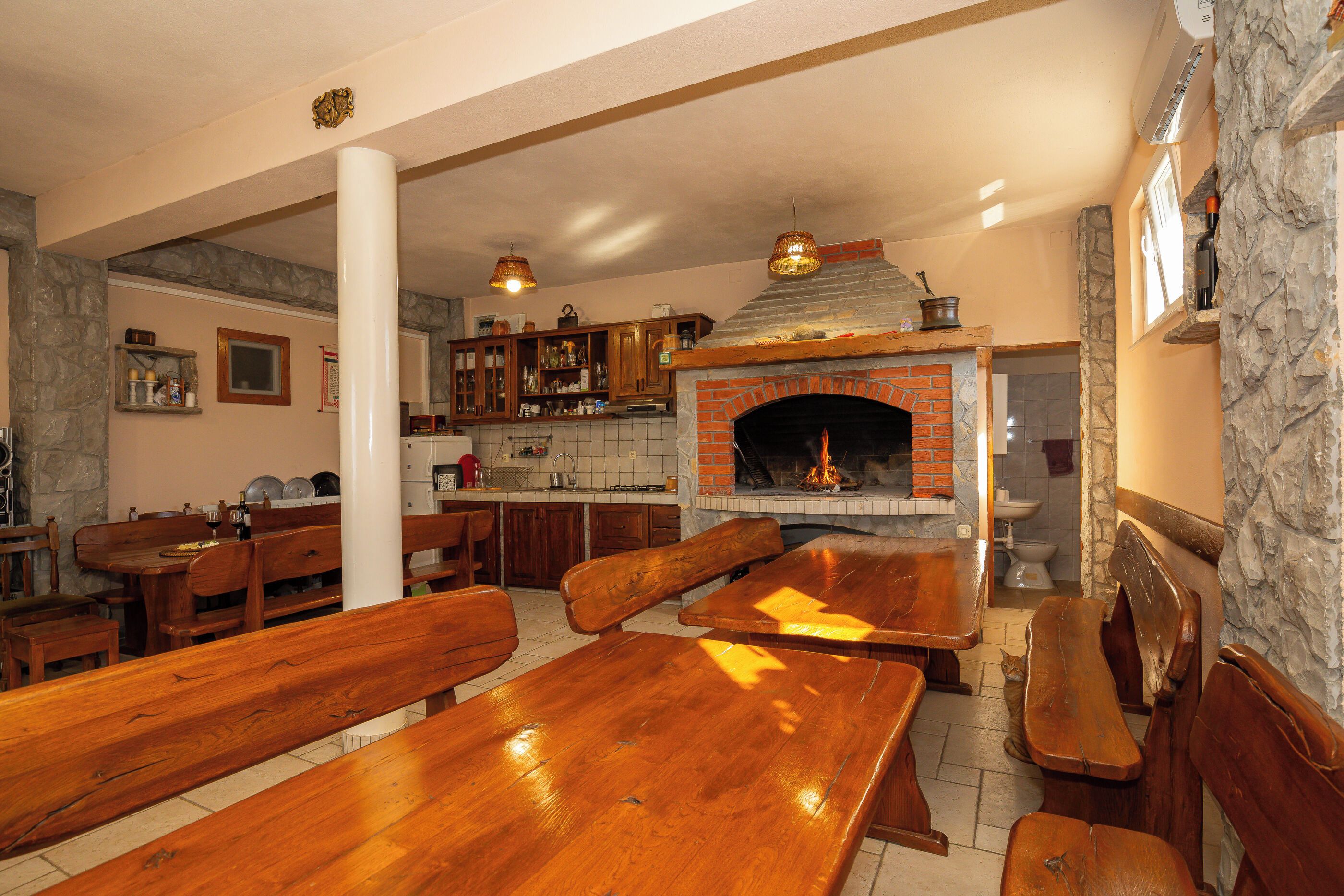 Ein Restaurant mit Holztischen und -bänken, einem Backsteinofen, Steinwänden und einer Küchenzeile mit Schränken und einer Hängelampe.