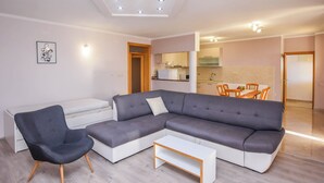 A modern living room with a grey sectional sofa, a small white coffee table, and a black armchair. There is a dining area with a wooden table and chairs visible in the background. - Apartments Svarog (Biograd)