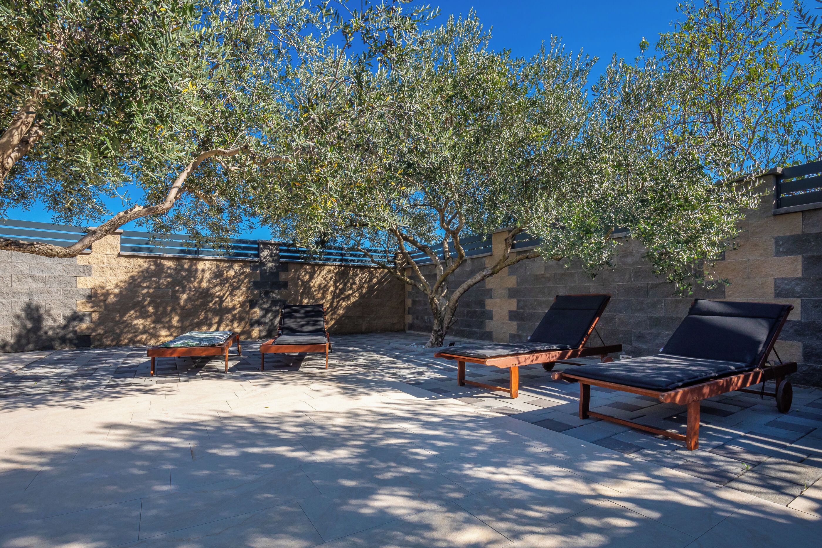 An outdoor seating area with lounge chairs and a small table, surrounded by stone walls and olive trees.