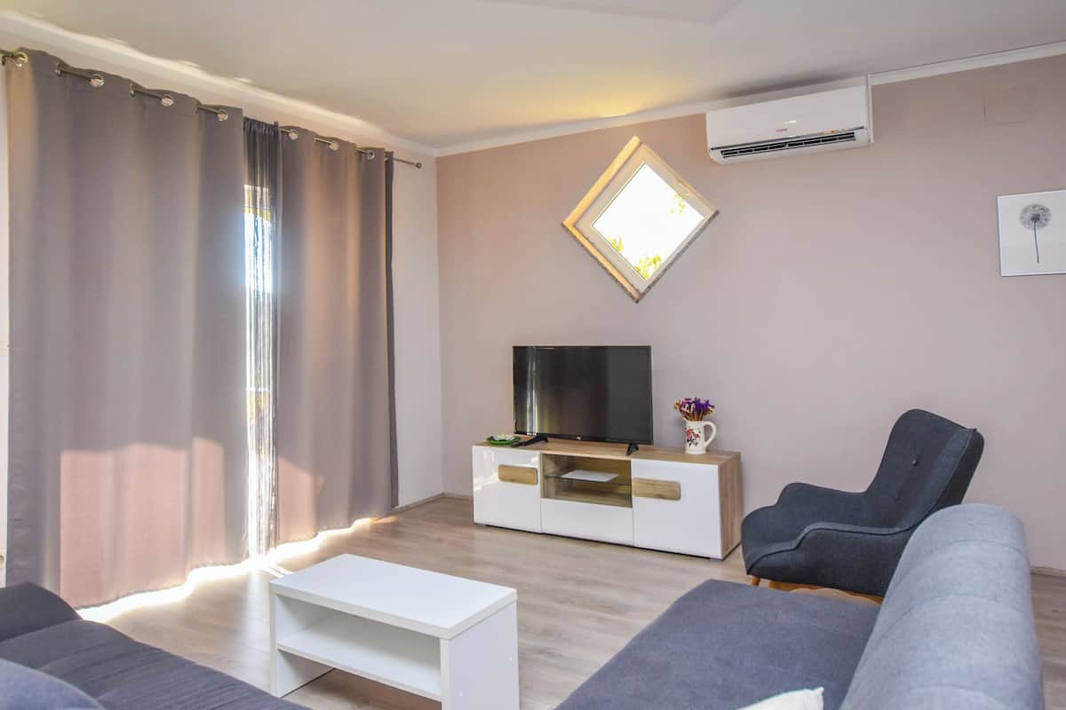 Un salon moderne avec un téléviseur à écran plat posé sur un meuble en verre, des canapés gris, une table basse blanche et une grande fenêtre avec des rideaux.