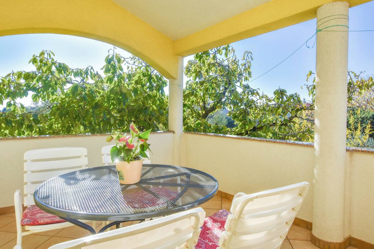 Une terrasse couverte avec une table ronde et des chaises blanches, un pot de fleurs et une vue sur les arbres.