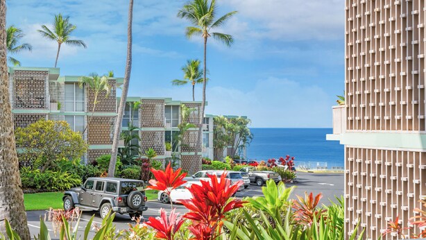 A hotel with a view of the ocean, palm trees, and a parking lot with vehicles. - 🌴 Perfect Island getaway | AC | Ocean View | AV #124🌴 (Kailua Kona)