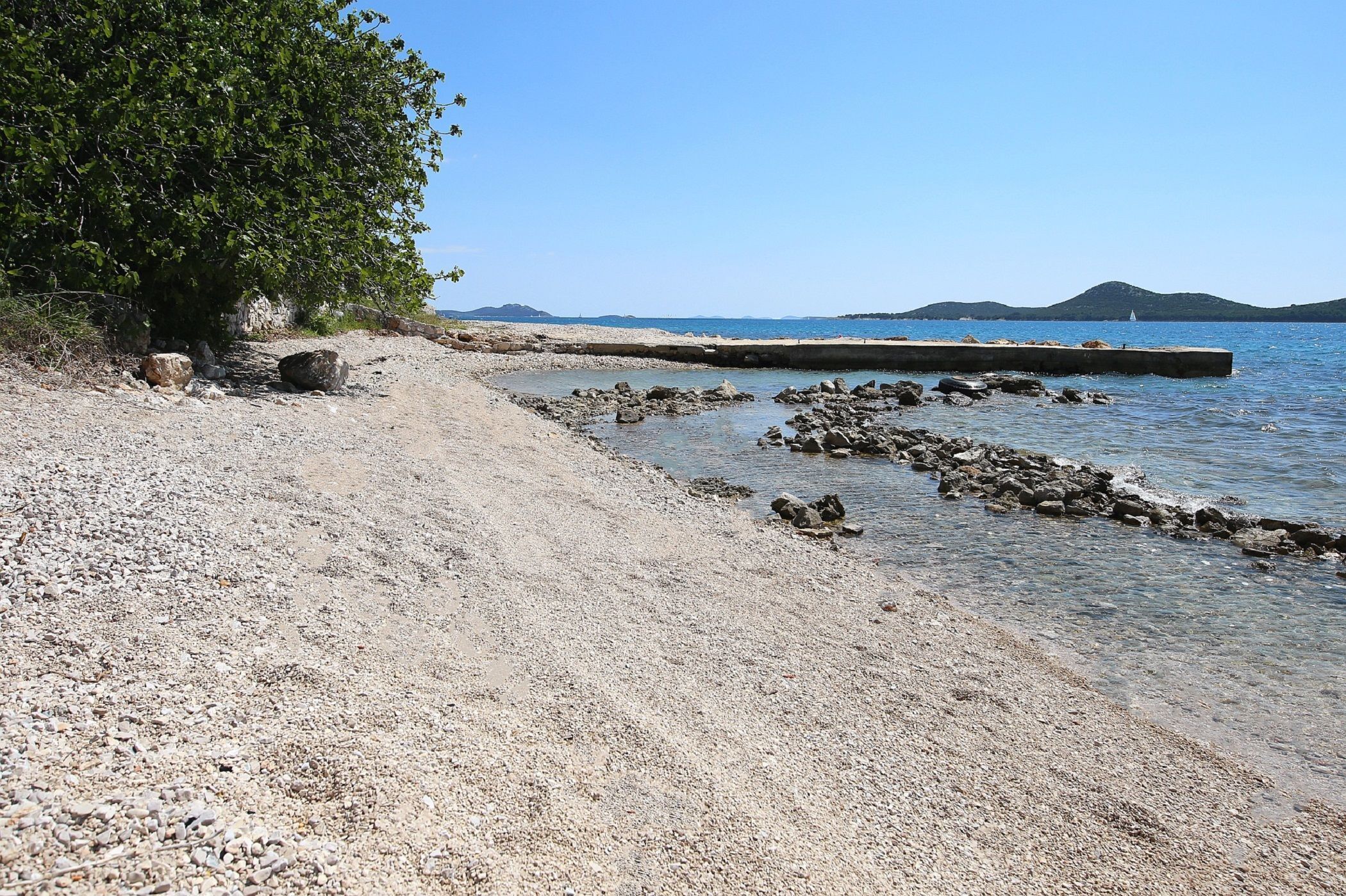 Una spiaggia con ciottoli, un albero e un molo roccioso che si protende verso il mare.