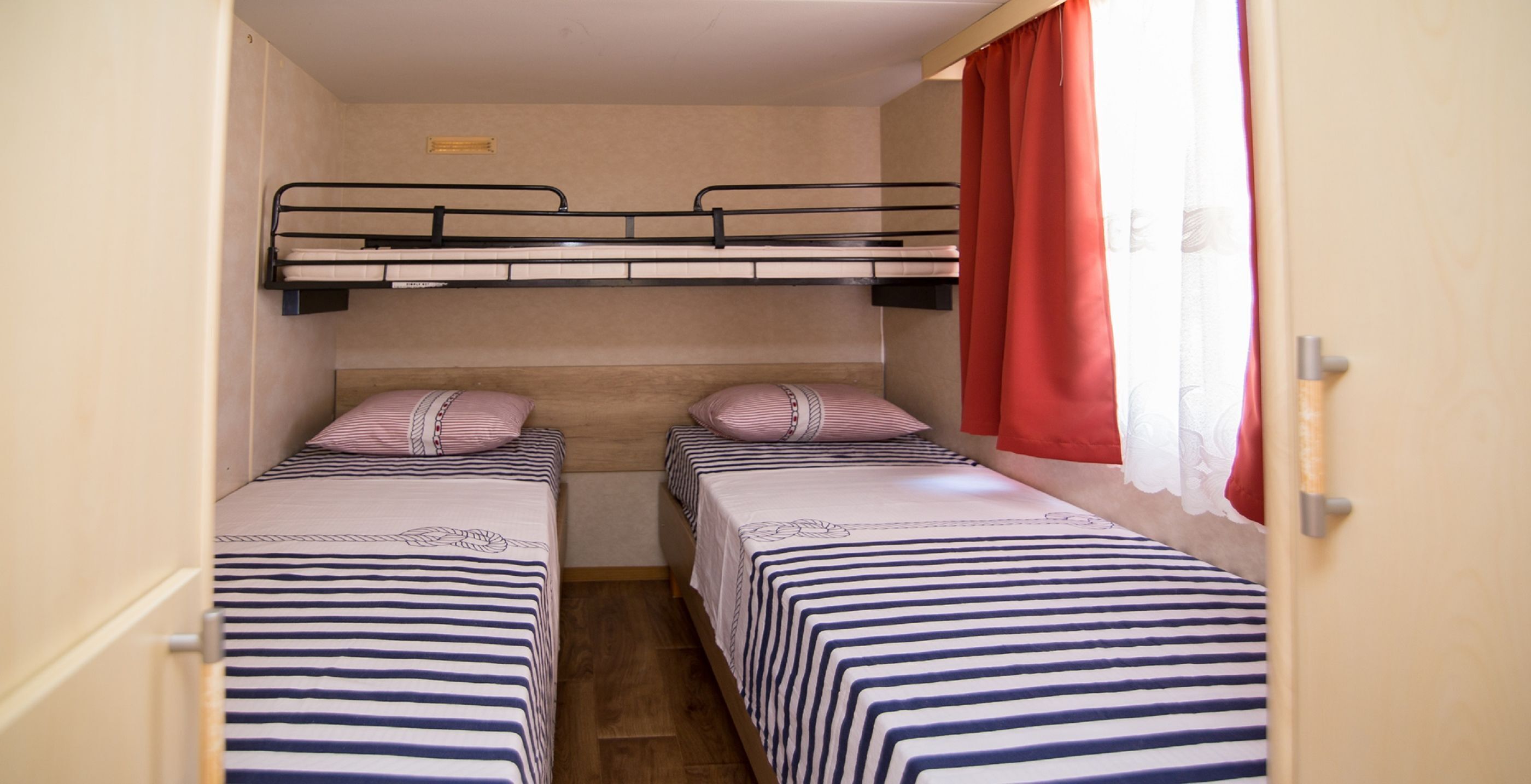 A compact room with two single beds, a bunk bed above, and a window with red curtains.