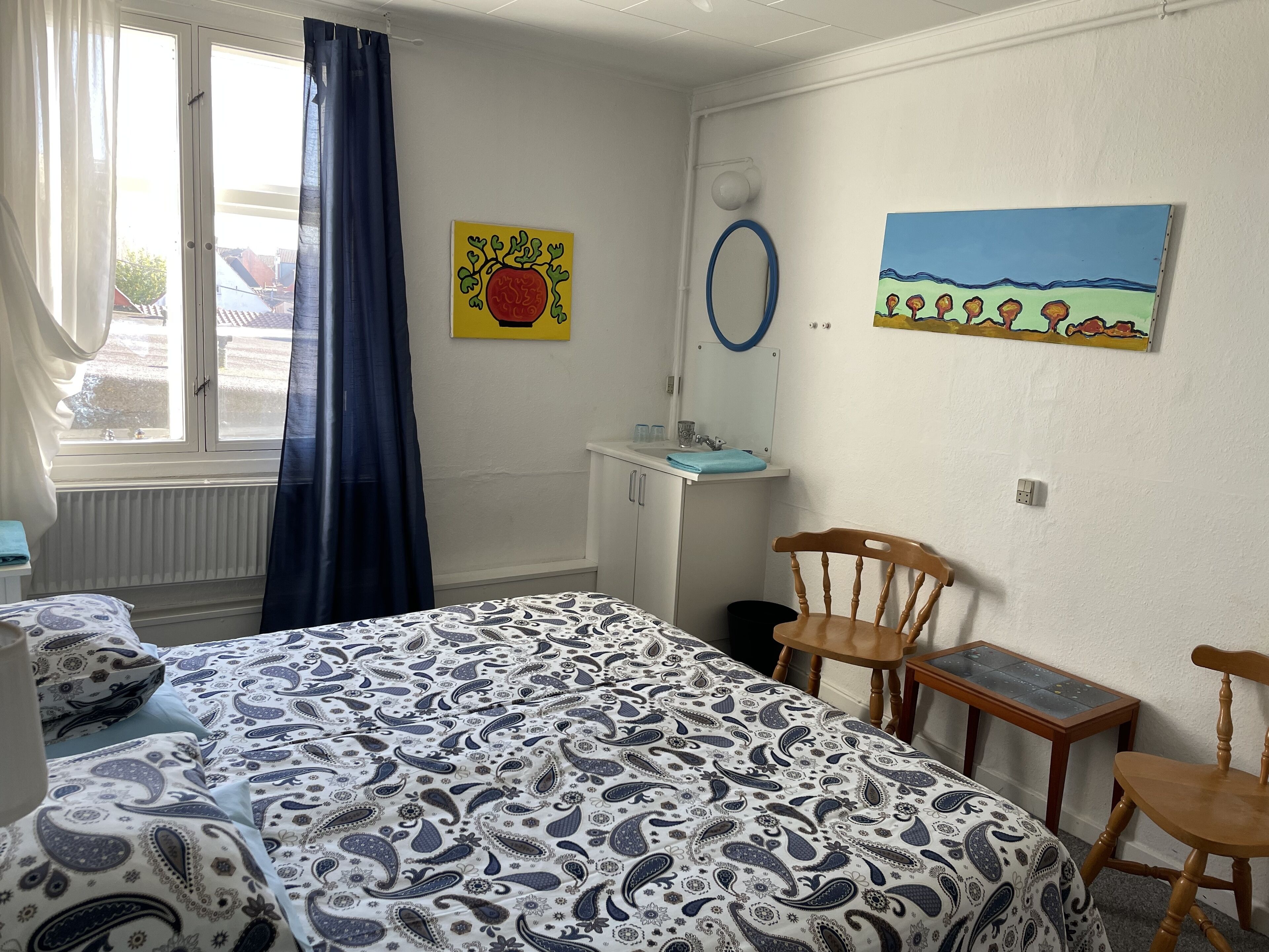 Un dormitorio con cama, escritorio, silla, ventana con cortinas y dos cuadros en la pared.