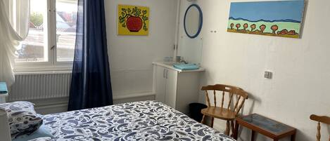 Un dormitorio con cama, escritorio, silla, ventana con cortinas y dos cuadros en la pared.