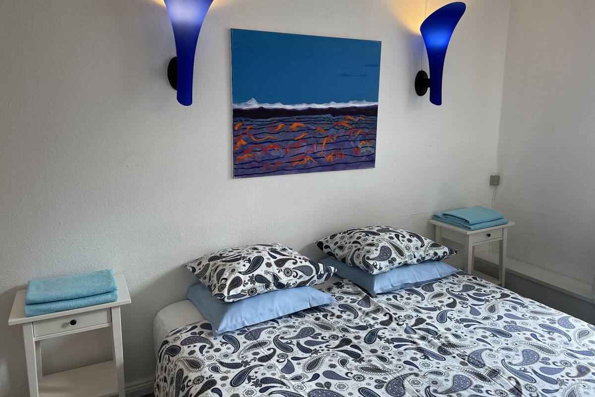 Quarto com cama, duas mesas de cabeceira, um quadro pendurado na parede e duas arandelas.