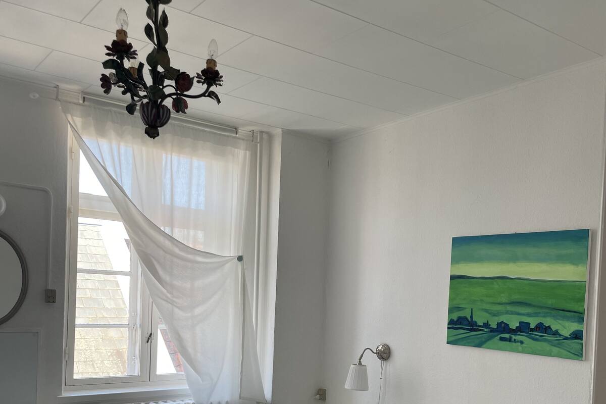 Um quarto com cama, mesa de cabeceira, luminária, janela com cortinas, radiador, quadro na parede e ventilador de teto.