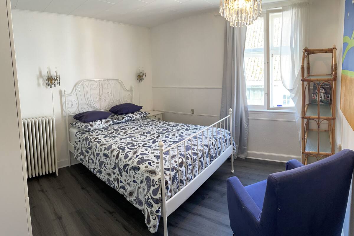 Quarto com cama, cadeira, radiador, lustre e janela com cortinas.