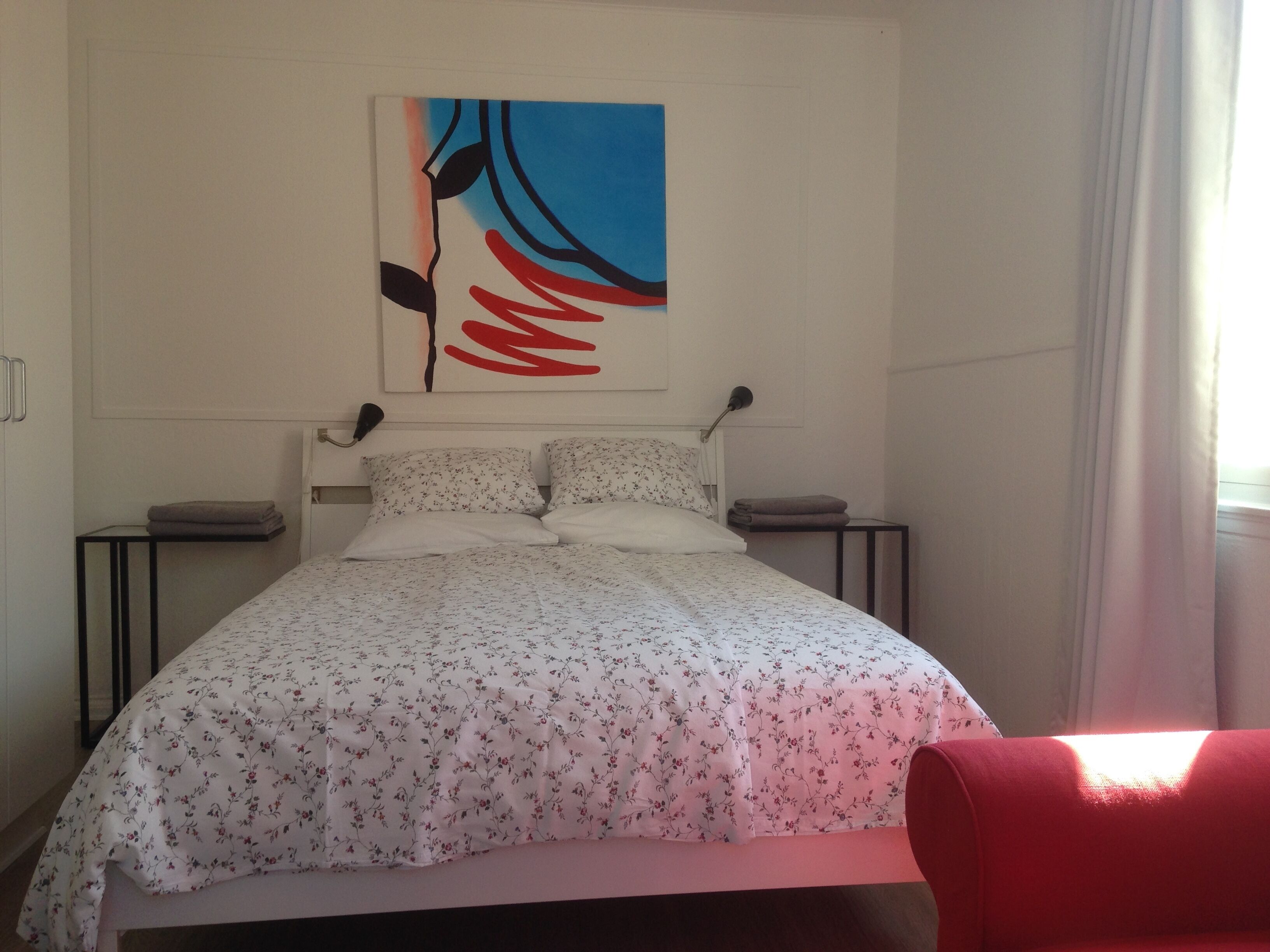 Una cama bien tendida con ropa de cama floral, una obra de arte colgada en la pared, lámparas de noche y una silla roja.