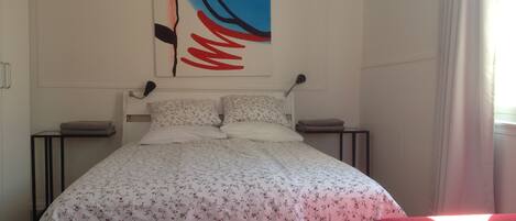 Una cama bien tendida con ropa de cama floral, una obra de arte colgada en la pared, lámparas de noche y una silla roja.