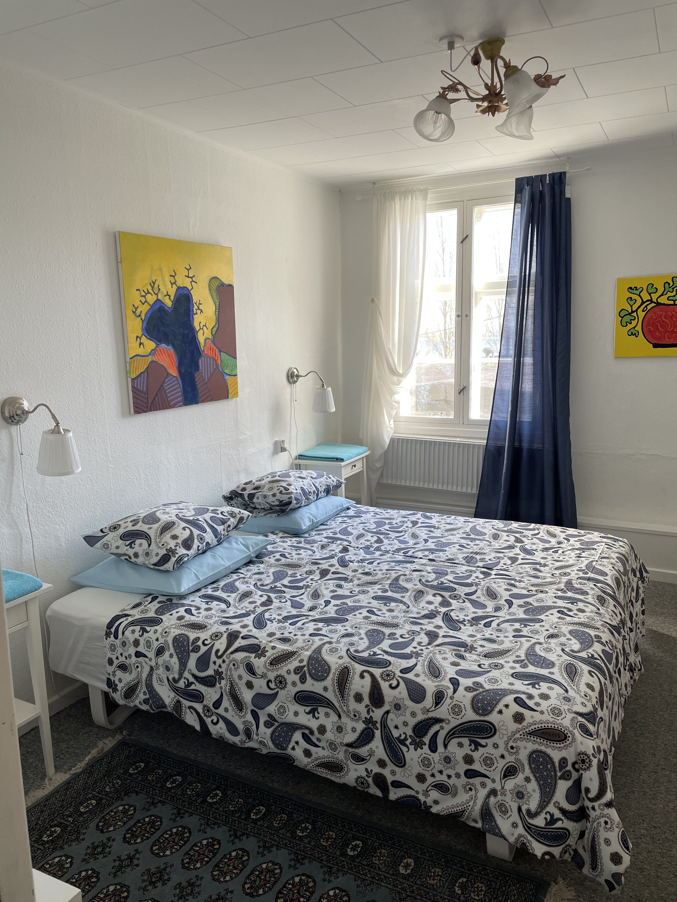 A bedroom with a bed, two bedside tables, a window with curtains, and a ceiling light fixture.