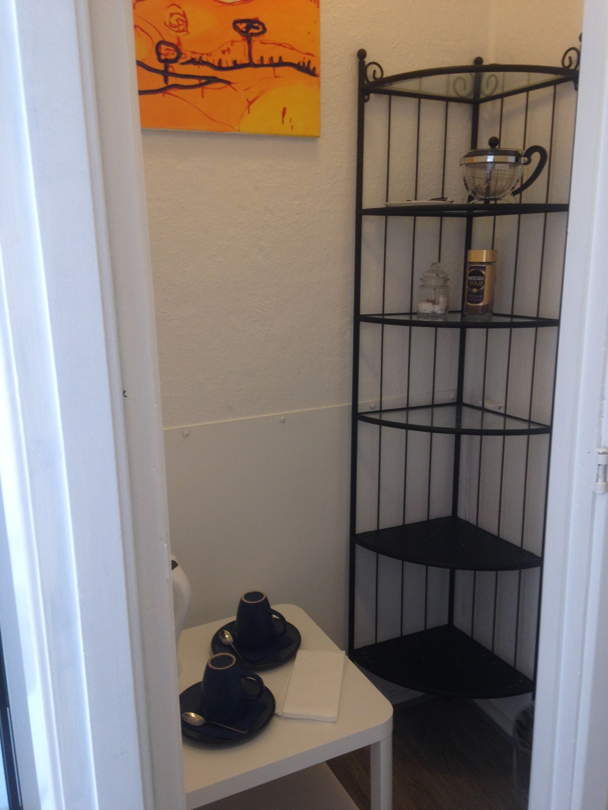 A corner with a black metal shelf unit, a white table with two cups and a spoon, and a painting on the wall.