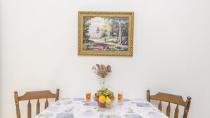 A dining table with a floral tablecloth, a fruit bowl, and a vase with dried flowers. A framed painting of a landscape hangs above the table. - Apartments Mira I (Vodice)