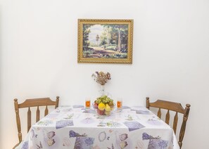 A dining table with a floral tablecloth, a fruit bowl, and a vase with dried flowers. A framed painting of a landscape hangs above the table.