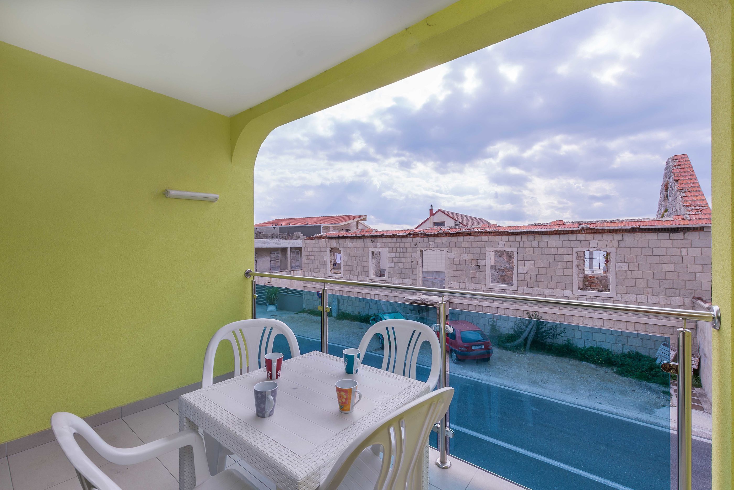 A balcony with a table and chairs, a view of a street with buildings and a car, and a cloudy sky.