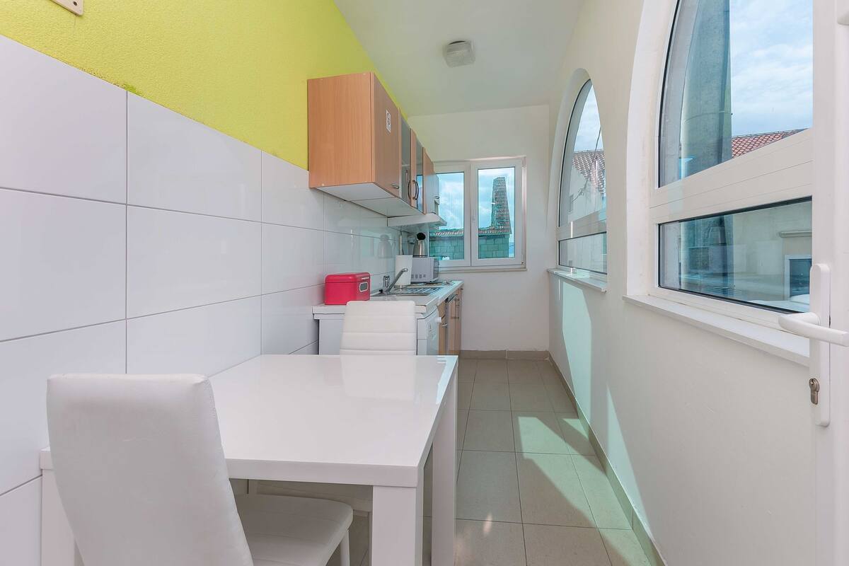 A narrow kitchen with white cabinets, a white table, and a window with a view of a building.