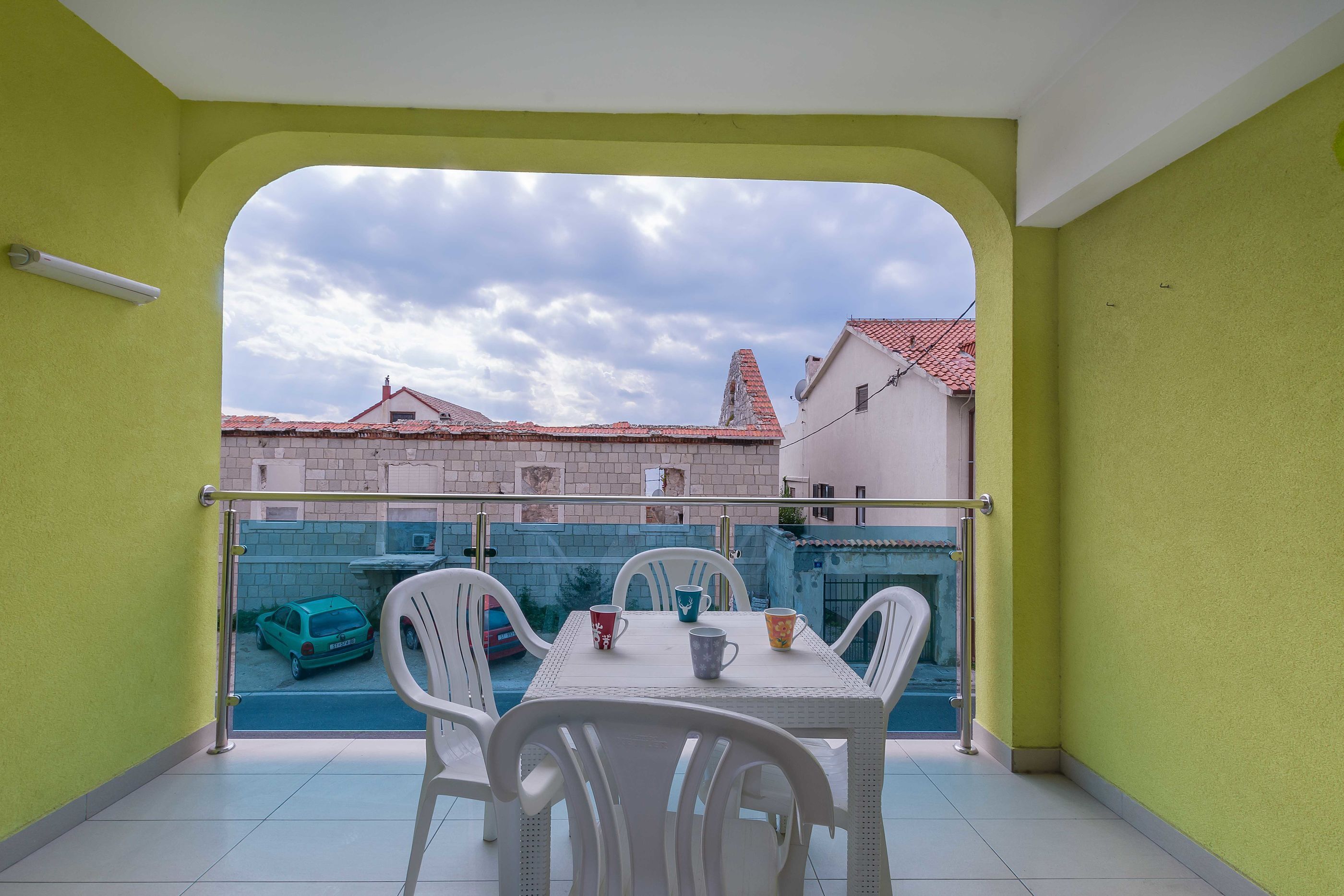 Un balcon avec une table et des chaises, une vue sur une rue bordée d’immeubles et un ciel nuageux.