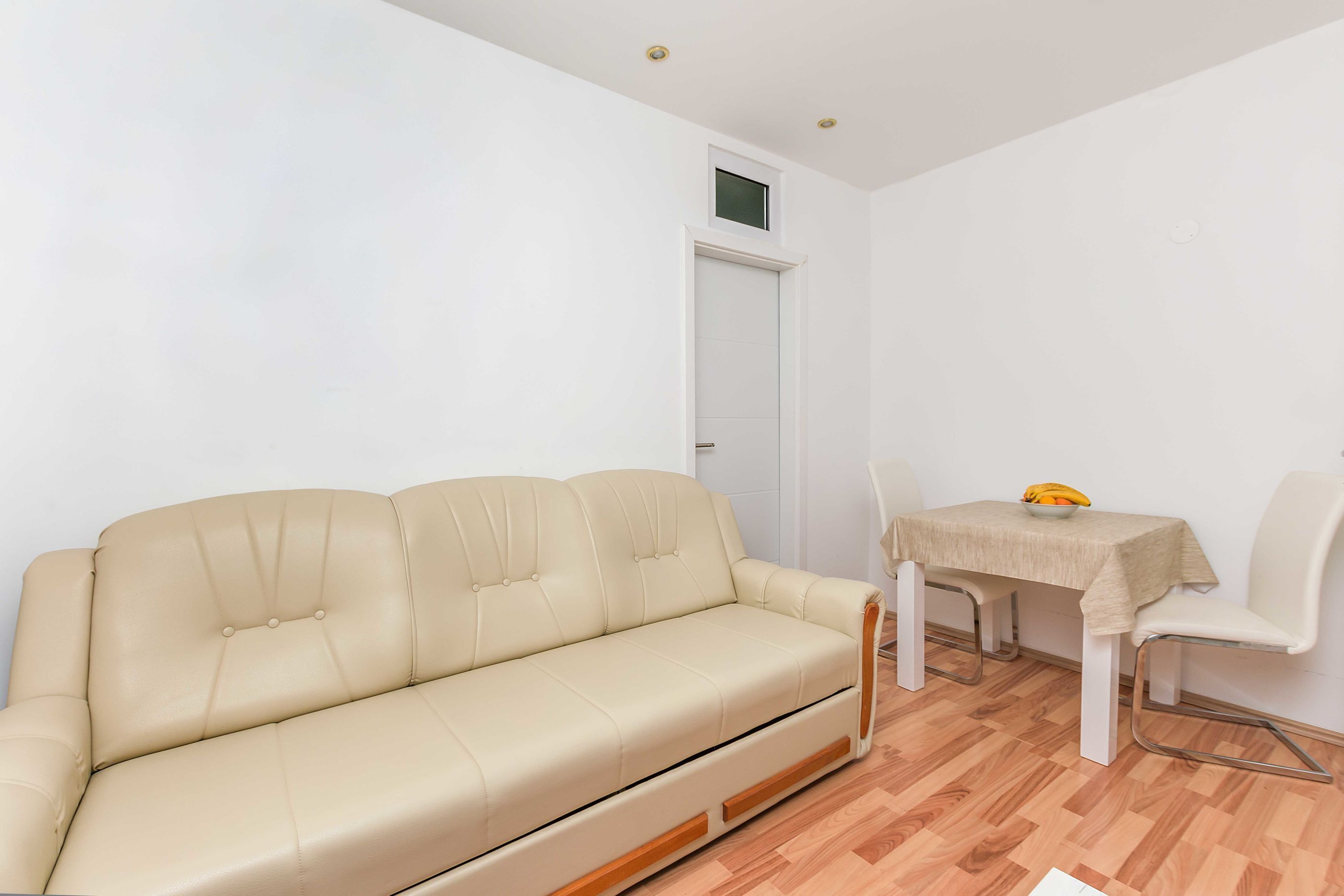 A room with a beige sofa, a dining table with a bowl of fruit, and a white door.