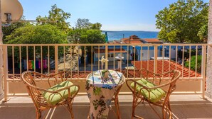 Un balcon avec des fauteuils en osier et une table, donnant sur une zone côtière avec des bâtiments et des arbres.