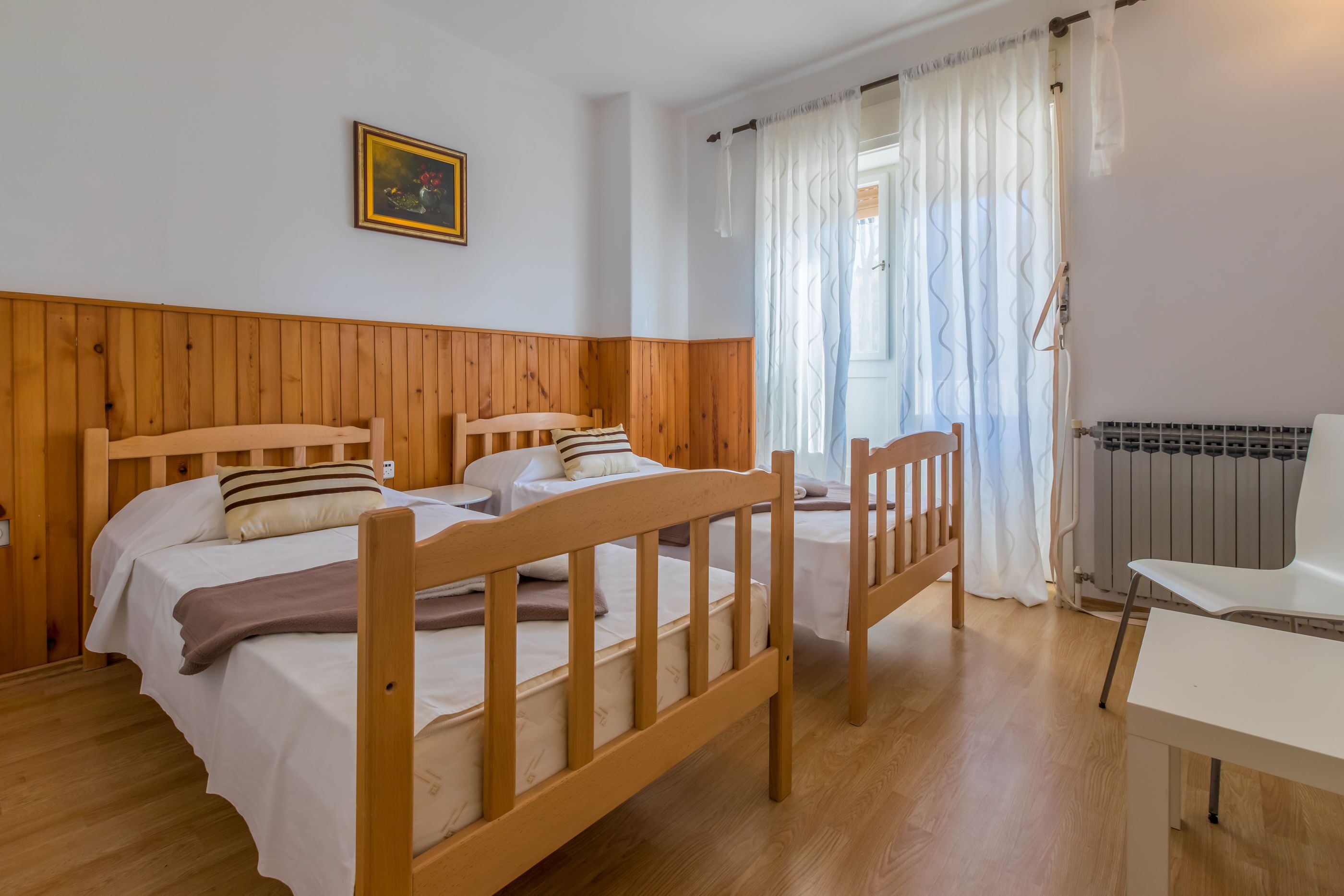 A room with two wooden beds, a white chair, a radiator, and a window with curtains.