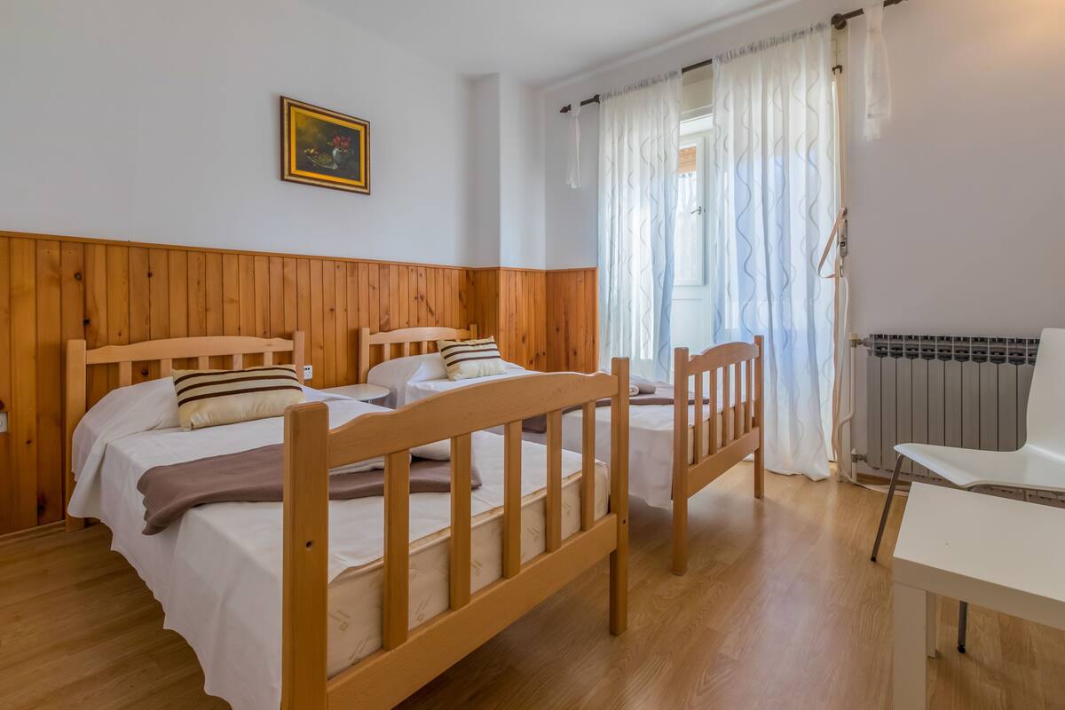 Egy szoba, amelyben két faágy, egy fehér szék, egy radiátor és egy függönnyel ellátott ablak található.