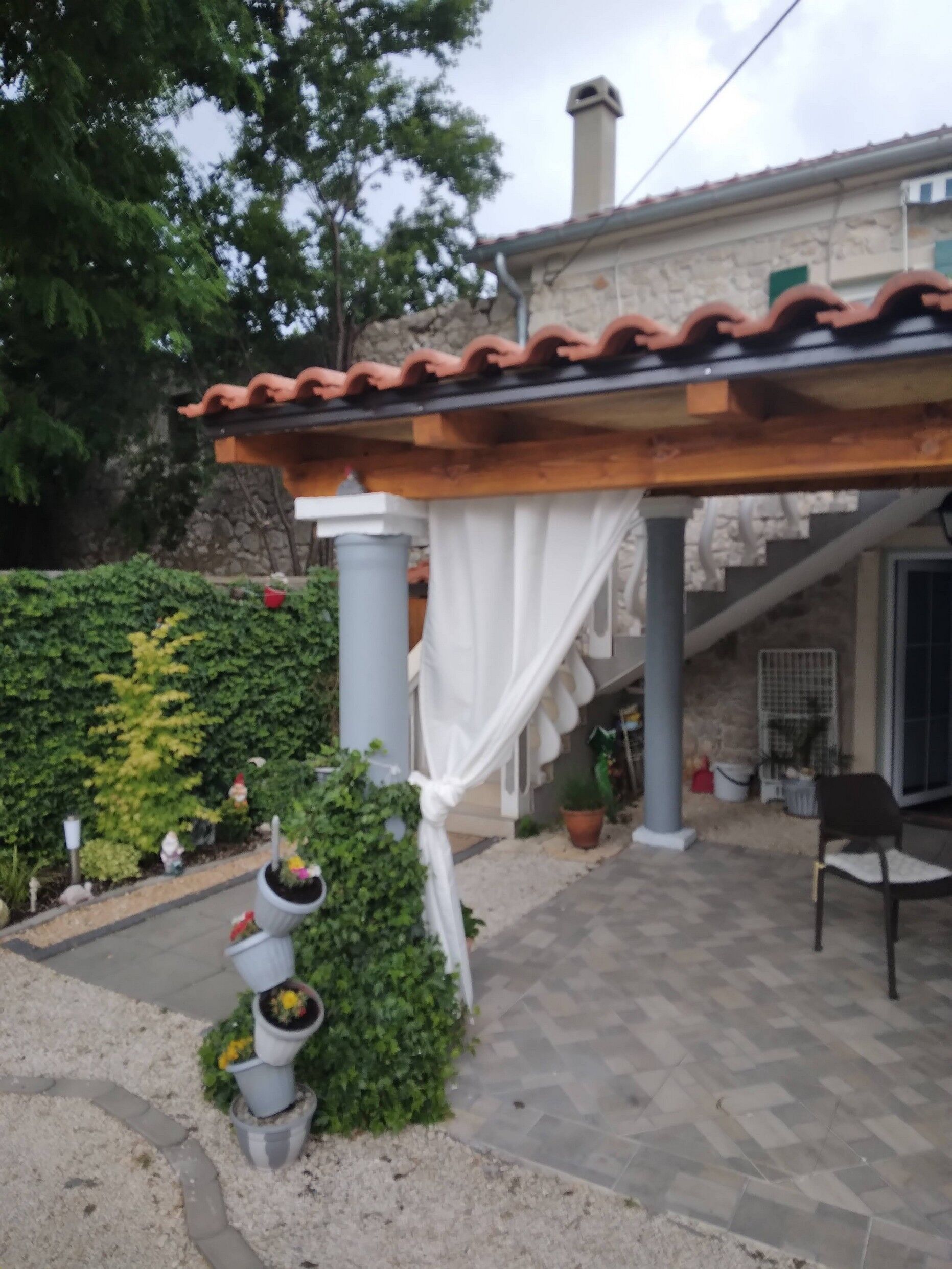 Un espace extérieur abrité, doté d’un sol carrelé, d’une pergola avec des rideaux blancs et de plantes en pot.