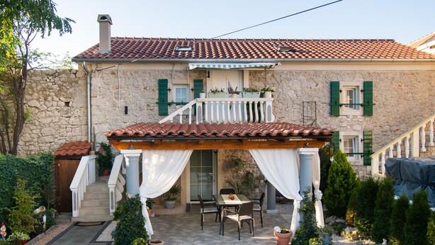 Ein Steinhaus mit Ziegeldach, eine überdachte Terrasse mit Tisch und Stühlen sowie ein kleiner Garten mit Topfpflanzen.