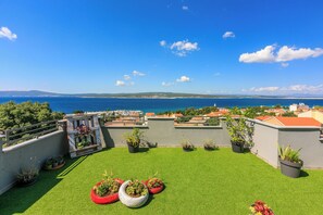 A rooftop terrace with artificial grass, potted plants, and a view of the sea and distant hills.