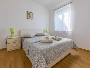 A bedroom with a bed, two bedside tables, a nightstand, a lamp, a picture on the wall, and a window with sheer curtains. - Apartments Benny (Crikvenica)