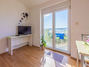 A room with a TV on a white stand, a sliding glass door to a balcony with a table and chairs, and a potted plant.