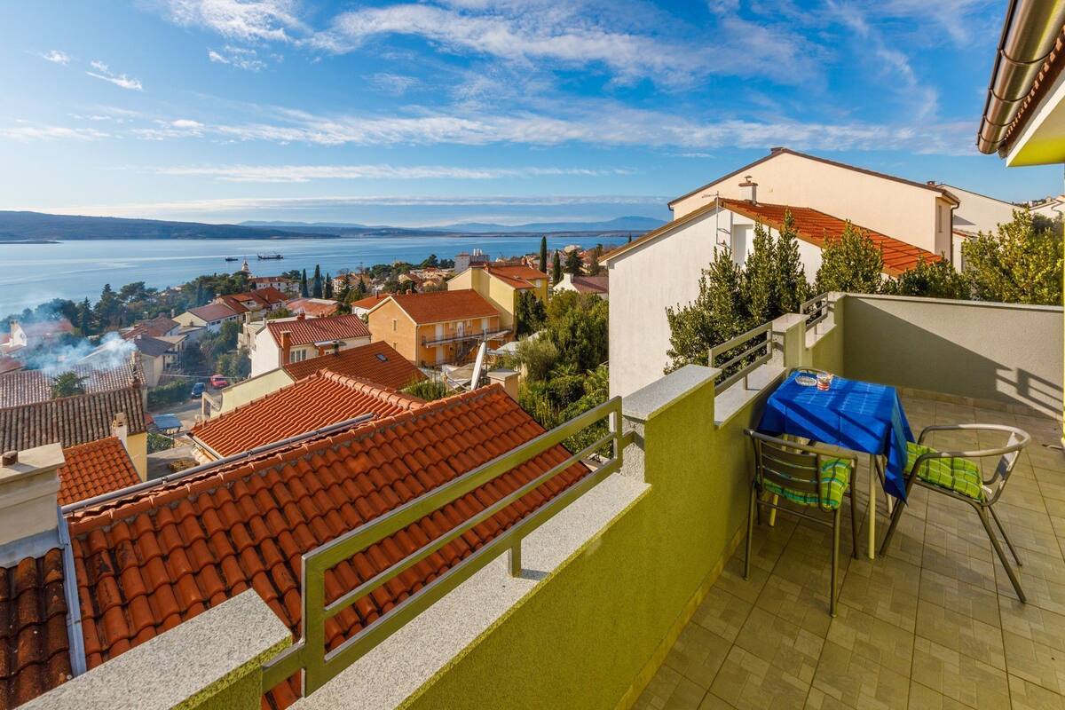 A balcony with a view of a coastal town, a table with chairs, and a railing.