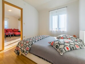 Een slaapkamer met een groot bed, een raam met dunne gordijnen en uitzicht op een zithoek met rode banken.