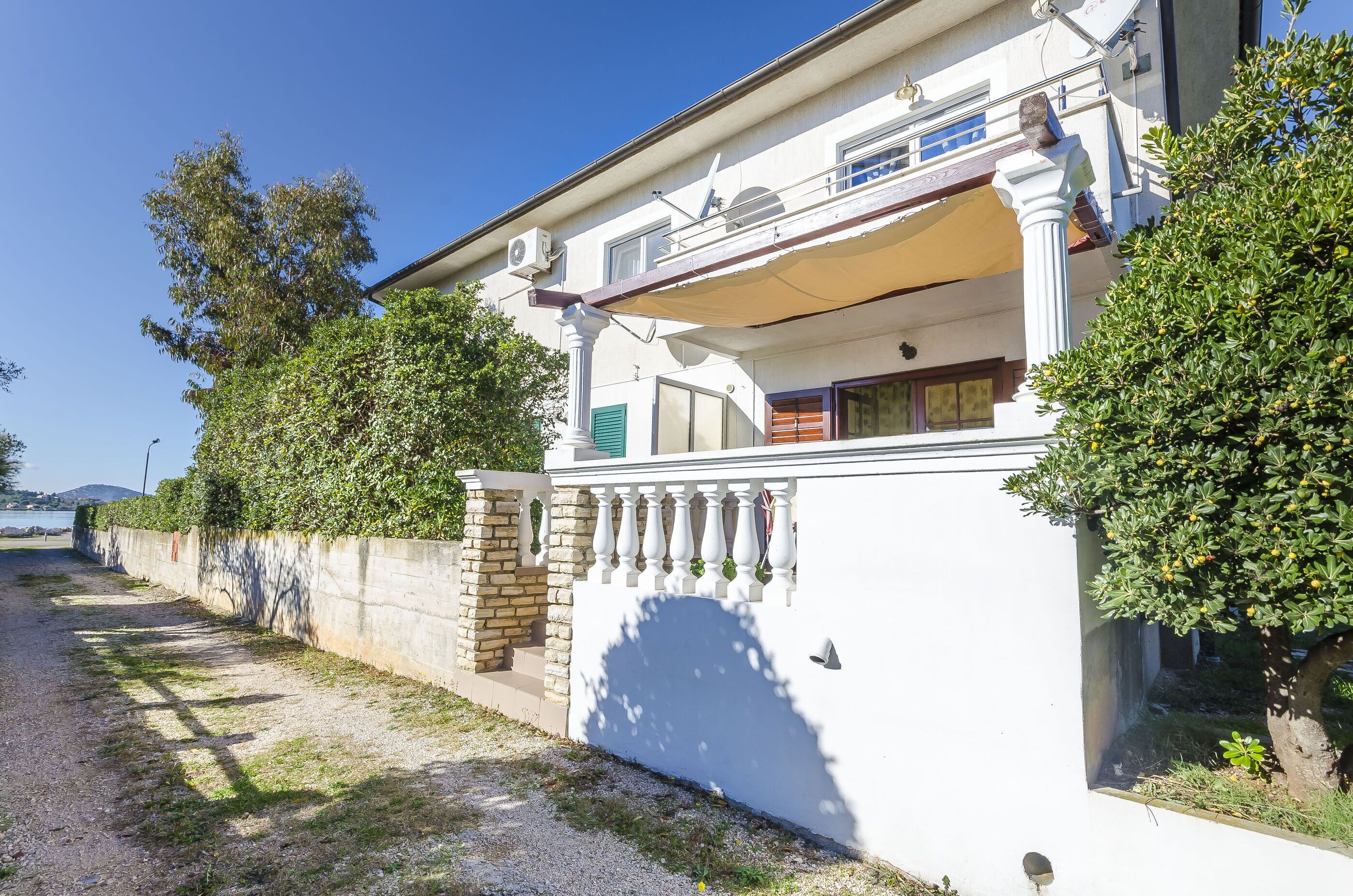 A white building with a balcony, a stone wall, and a tree.