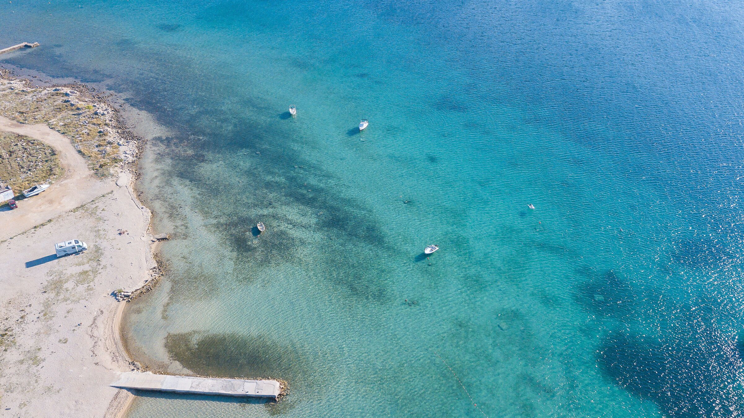 Vista aérea de una zona costera con aguas turquesas cristalinas, barcos y una playa arenosa con un espigón.