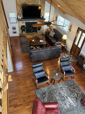 A spacious living room with a stone fireplace, a large flat-screen TV, a ceiling fan, and a cozy seating area with sofas and chairs.