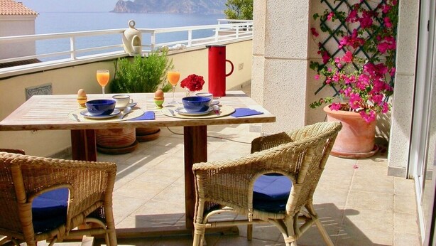 Un balcón con mesa dispuesta para el desayuno, sillas de mimbre, una planta en maceta y vistas al mar y a la montaña.
